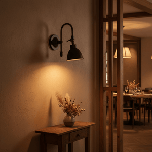 Matt black wall lamp with a curved arm illuminating a rustic wooden table with a vase of dried flowers.