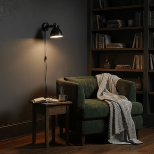 Maxim Cover Wall Lamp in matte black mounted above a green armchair with a cozy blanket and a coffee cup on a side table.