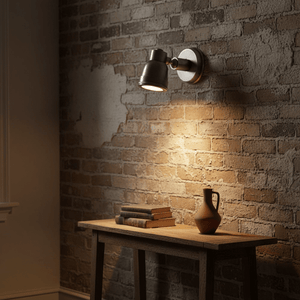 Indigo Wall Lamp Swirl in antique silver mounted on a textured brick wall, illuminating a wooden table with books and a vase.