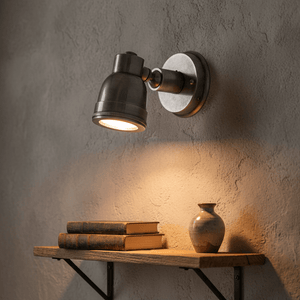 Indigo Wall Lamp Swirl in antique silver mounted on a textured wall, illuminating a wooden shelf with books and a vase.
