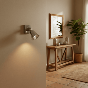 Antique silver wall lamp illuminating a textured wall, with a wooden table and plants in the background.