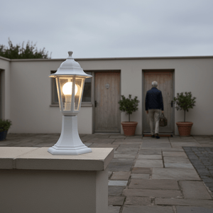 White six-panel pedestal lantern illuminating a pathway near two wooden doors and potted plants.