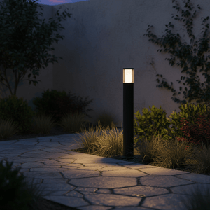 A sleek black LED bollard illuminates a stone pathway surrounded by lush greenery at dusk.