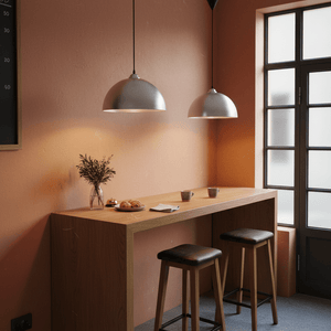 Two aluminium pendant lights hang above a wooden counter with stools, cups, croissants and a vase against a peach wall.