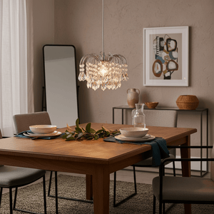 Polished chrome pendant light with clear acrylic crystals hanging over a wooden dining table set with plates and greenery.