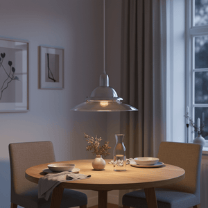 Silver aluminium pendant light with clear glass shade hangs over a round wooden table set with dishes and a vase.