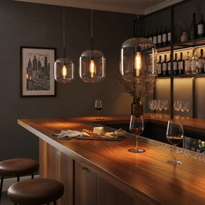 Three metal pendant lights with cognac glass shades hang above a wooden bar, illuminating wine glasses and a cheese platter.
