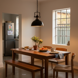 A black and gold metal pendant light hangs over a wooden dining table set with plants and fruit.