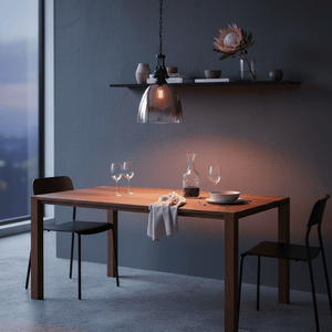 Black metal pendant light with faded rose gold glass shade hanging over a wooden dining table set for two.