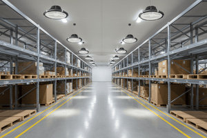 LED high bay lights illuminate a warehouse aisle filled with stacked boxes on pallets.