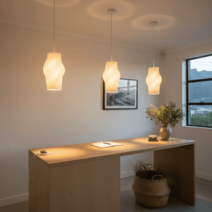 Three white pendant lights hang above a wooden table, illuminating a modern indoor space with a plant and artwork.