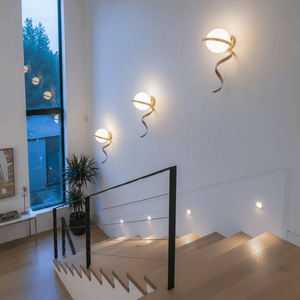Three gold infinity wall lights mounted on a white wall beside a staircase, illuminating the space with a warm glow.