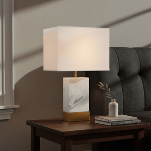 Bach Gold & Marble Table Lamp on a wooden side table beside a grey upholstered sofa, casting warm light.