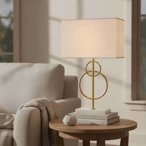 Beethoven Gold & Marble Table Lamp on a wooden side table beside a cozy sofa, with books and a decorative bowl.