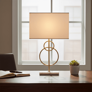 Elegant table lamp with a gold base and fabric shade on a wooden desk beside a notebook and plant.