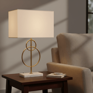 Elegant table lamp with a gold base and fabric shade on a wooden side table beside a beige armchair.
