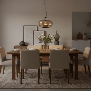 A modern dining room setup featuring a rectangular wooden dining table with six upholstered chairs in a neutral beige tone, under a warm amber glass pendant light. The room has a beige wall, a patterned area rug, and a glimpse of a kitchen in the background.