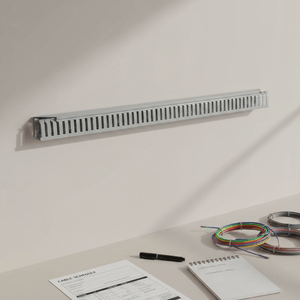 A long, narrow grey plastic cable management tray mounted on a light beige wall above a desk. The desk includes papers, a black pen, and coiled multicolored cable ties.