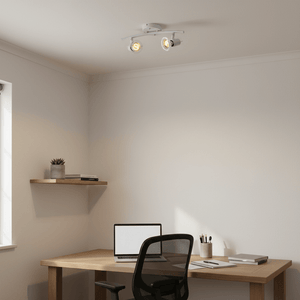 White two-light indoor spotlight mounted on ceiling above a wooden desk with a laptop and stationery.