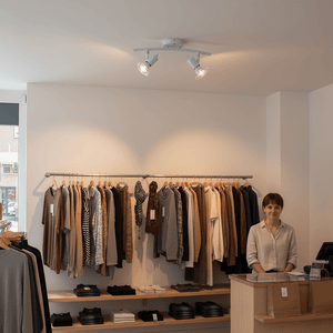 White two-light indoor spotlight illuminating a retail space with clothing displays and a cashier.
