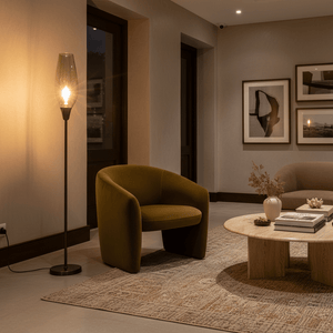 Amber glass floor lamp with black metal stand, illuminated, standing beside a green armchair in a living room.