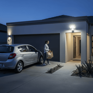 A woman carrying groceries approaches a well-lit entrance, with a motion sensor floodlight illuminating the driveway.