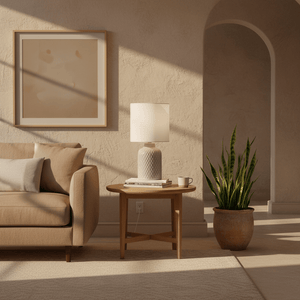 A stylish Castille Table Lamp on a wooden side table next to a beige sofa, with a plant and artwork in the background.