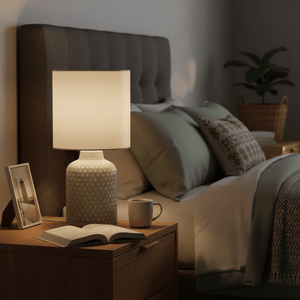 A stylish ceramic table lamp on a bedside table, illuminating an open book and a coffee mug in a cozy bedroom setting.