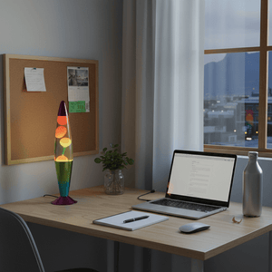 Colorful lava lamp on a desk beside a laptop, with a corkboard and plant in the background.