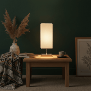Satin chrome table lamp with beige shade on a wooden table, surrounded by books, a cup, and a decorative vase.