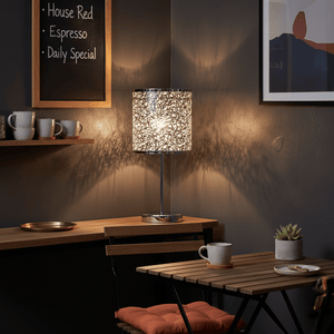 Polished chrome table lamp with a silver patterned shade casting intricate shadows in a cozy indoor setting.