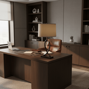 A black and gold table lamp sits on a dark wood desk, surrounded by books, a laptop, and stationery in a modern office setting.