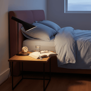Black metal table lamp with adjustable head illuminating a bedside table with a book, glasses, and a clock.