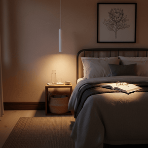A slim white pendant light hangs above a bedside table, illuminating an open book and glassware.