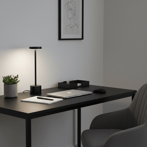 Modern black rechargeable table lamp on a sleek desk with a laptop, notebook, and plant.
