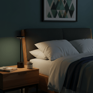 Vogue Black Rechargeable Table Lamp on a bedside table next to a cozy bed with pillows and a plant.