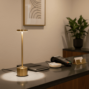 Gold rechargeable table lamp on a dark countertop, surrounded by a notebook, phone, and plant.