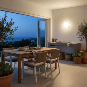 Round white coastal LED bulkhead wall light glowing warm on textured wall, with dining table and sea view