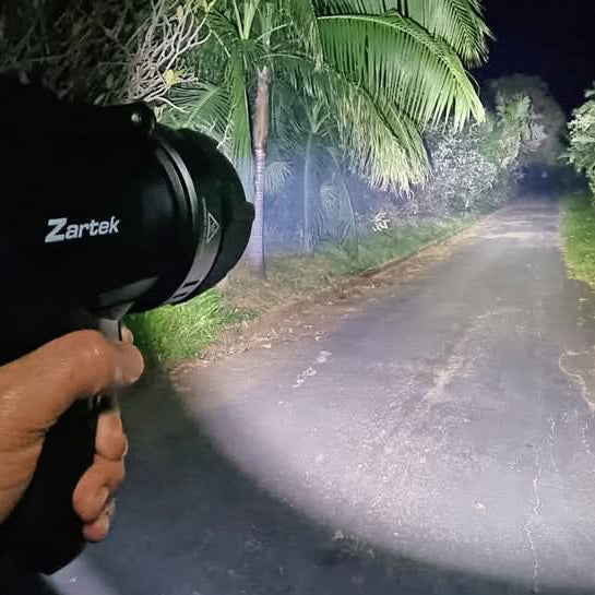 Handheld Zartek spotlight casting a focused white beam along a tree-lined road at night.