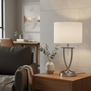 Satin chrome table lamp with oval pearl white shade on a wooden bedside table, complemented by a vase and artwork.