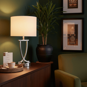Satin chrome table lamp with oval pearl white shade on a wooden surface, surrounded by decor and greenery.