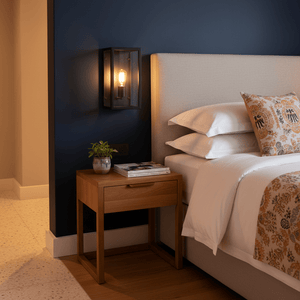 Metal indoor wall light with clear glass mounted beside a bed, illuminating a cozy bedroom setting.