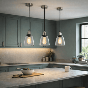 Three satin chrome pendant lights with clear glass shades hang over a kitchen island, illuminating a bowl of olives.