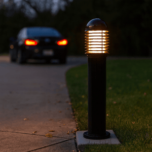 Black base mount bollard with louvre design illuminating a pathway at dusk, with a car in the background.