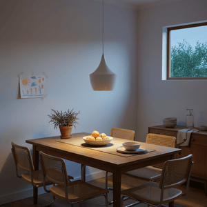 A contemporary white metal pendant light hangs above a wooden dining table set with fruit and tableware.
