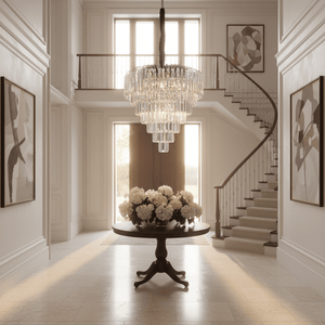 Crystal chandelier hangs over a dark round table with white hydrangeas in a bright double‑volume foyer with curved staircase.