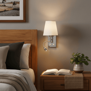Polished chrome wall light with white shade mounted beside a wooden headboard, illuminating a bedside table with a book.
