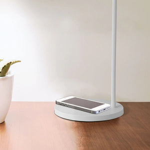 Close-up of a white desk lamp with a wireless charging base, smartphone placed on it, and a potted plant nearby.