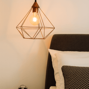Geometric pendant light above a bed with pillows and an alarm clock on a nightstand.