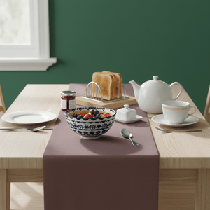 A mini ceramic bowl with orange and blue diamond patterns sits on a table with a mauve table runner, surrounded by dishes and a teapot.